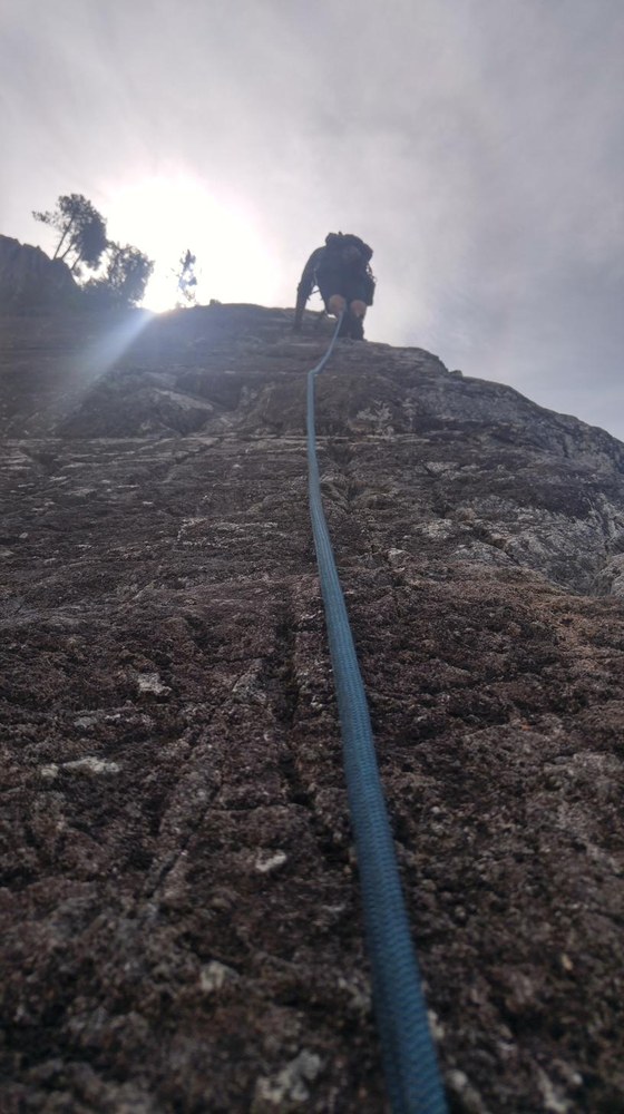 A climber ascending a steep rock face toward the sun, rope leading up from the foreground