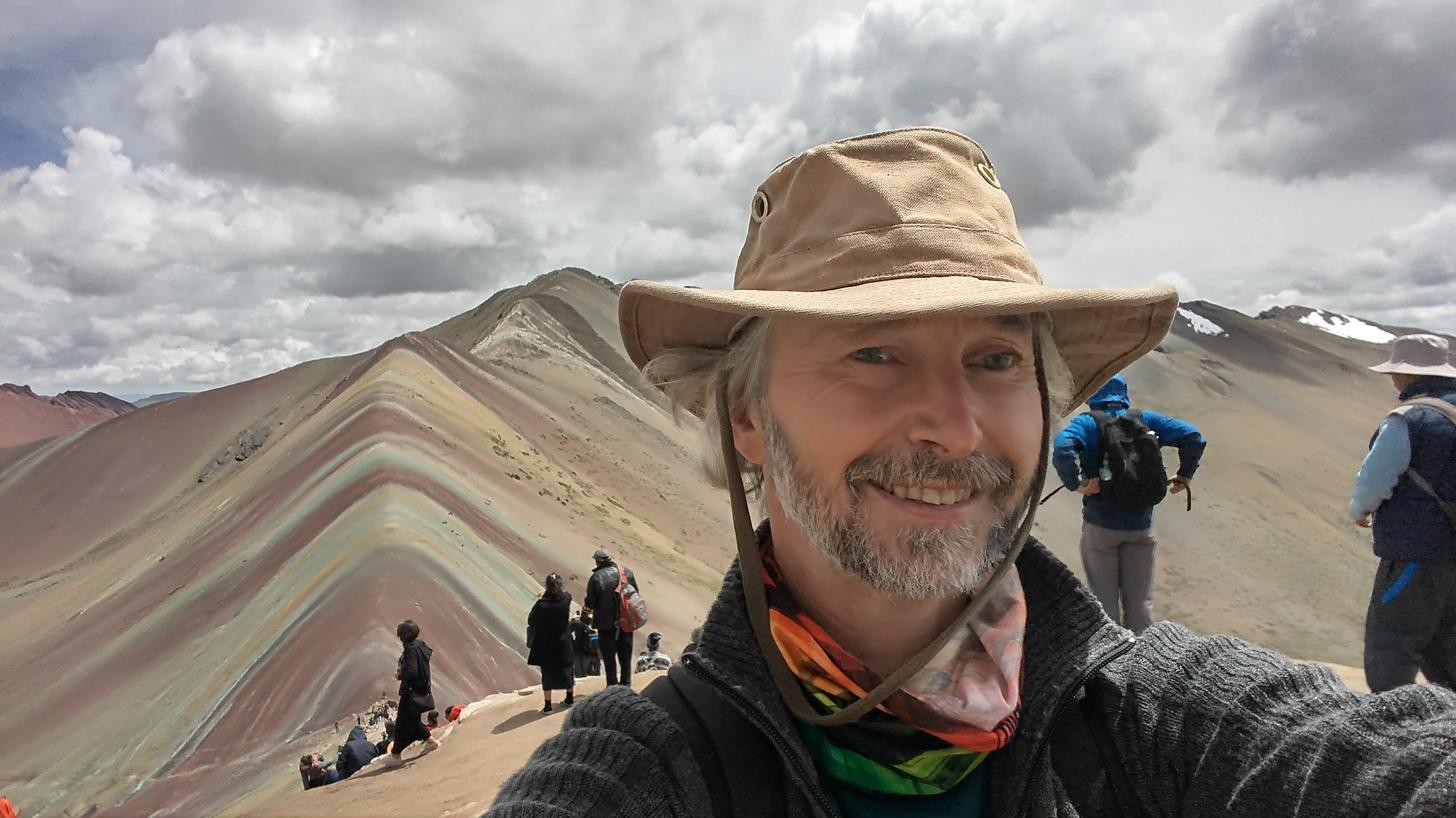 Todd at Rainbow Mountain Peru, layered terracotta and sage peaks behind him